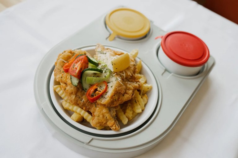 Knuspriges Schnitzel mit Pommes im Transporttablett, dazu zwei Saucenbehälter (rot und gelb).