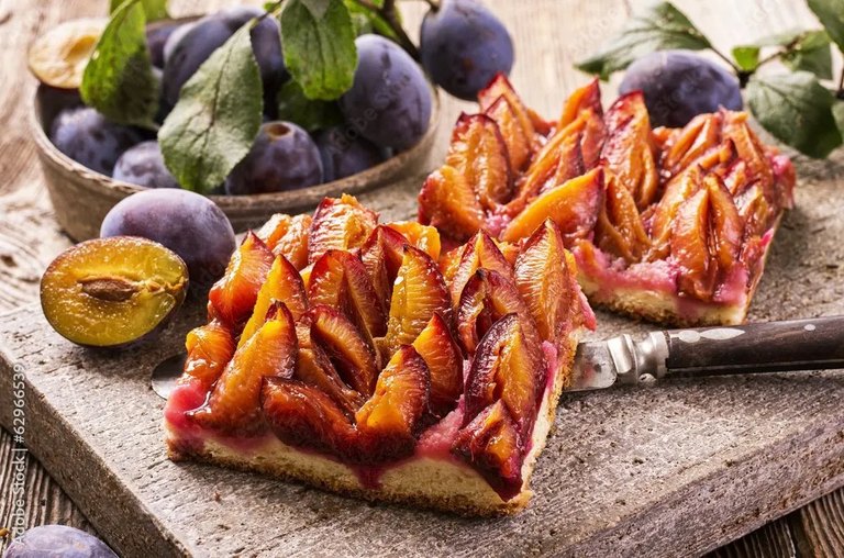 Zwei Stücke frischer Zwetschkenkuchen mit ganzen Zwetschken im Hintergrund auf einem Holzbrett.