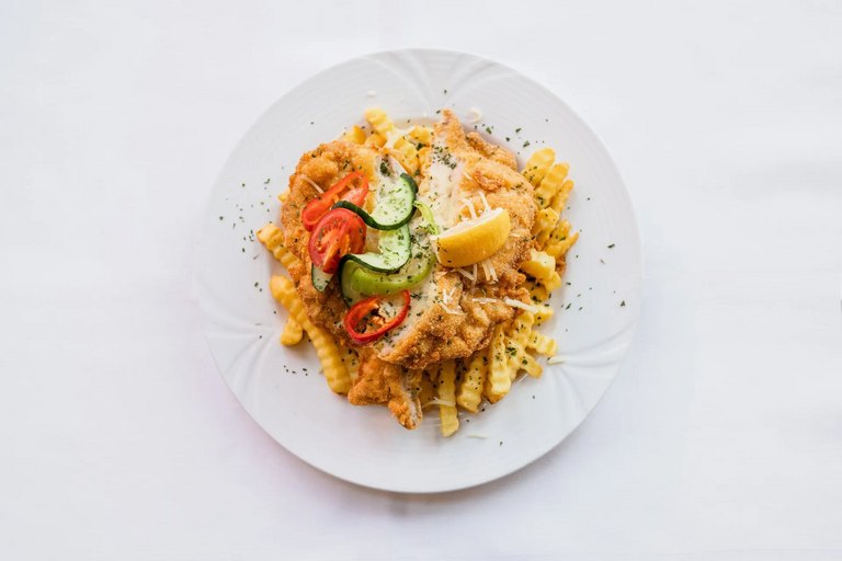 Knuspriges Schnitzel auf gewellten Pommes, garniert mit Gurken- und Tomatenscheiben sowie einer Zitronenspalte auf weißem Teller.