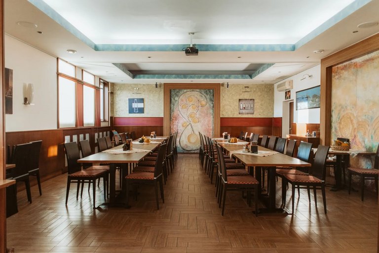 Großer Speisesaal mit zwei langen Holztischen, je Reihen von Stühlen und einem farbenfrohen Wandbild am Ende des Raums.