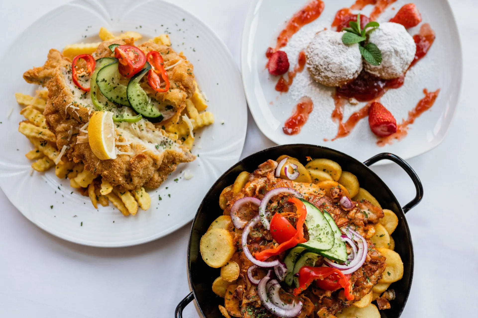 Weißer Teller mit Schnitzel auf Pommes, Puderzucker-Küchlein mit Erdbeersauce und Pfanne mit Bratkartoffeln und Gemüse