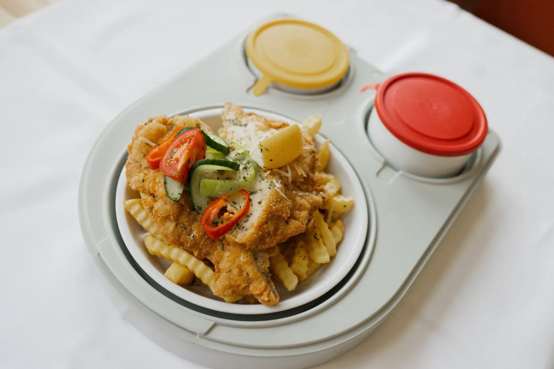 Knuspriges Schnitzel auf gewellten Pommes mit Gurke, Tomate, Zitrone und zwei Saucendosen auf einem Tablett