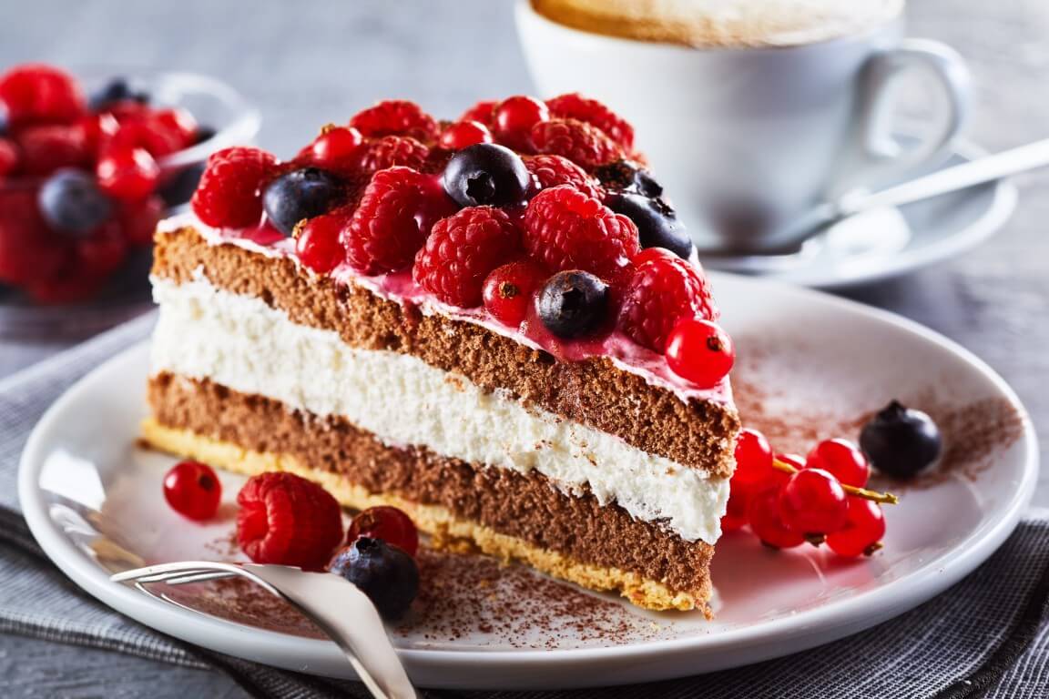 Stück Schokoladen-Sahnetorte mit Himbeeren und Johannisbeeren auf Tellerrand, im Hintergrund eine Tasse Kaffee